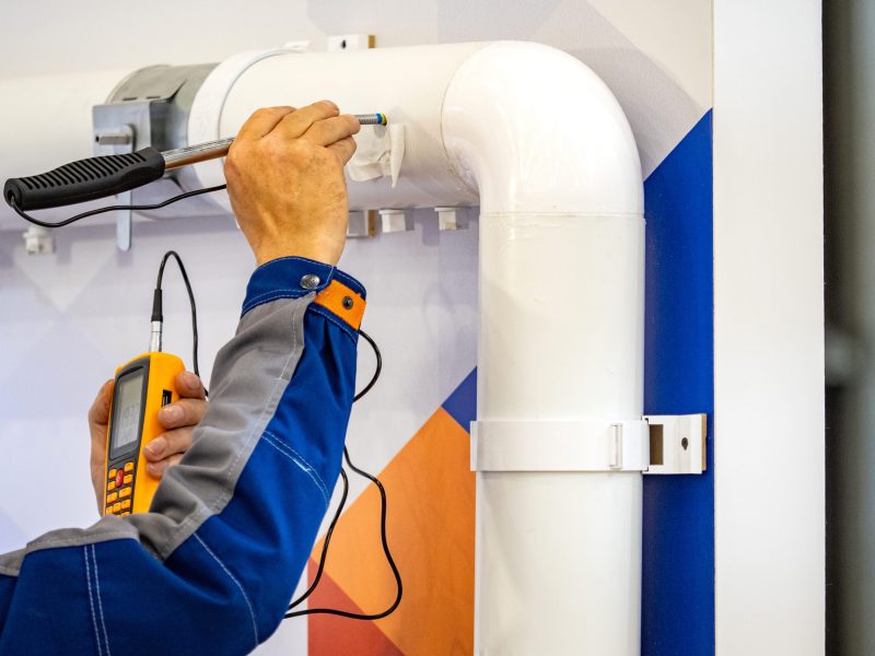 Installation of utility lines in building. Man uses scanner to find blockage in pipe. Craftsman stands near white pipeline fixed to wall. Process installation ventilation utility lines at enterprise